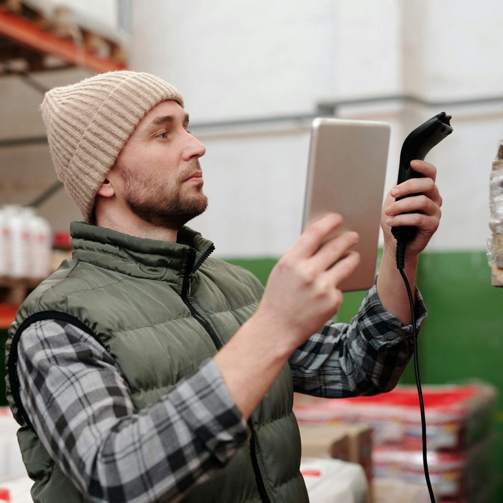 Man in beanie and vest using a scanner in a warehouse for inventory control.